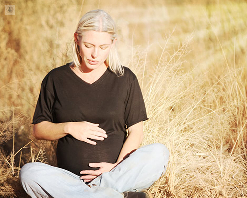 Chica embarazada sentada en un prado de trigo tocándose la tripa - Psicología y embarazo - by Top Doctors