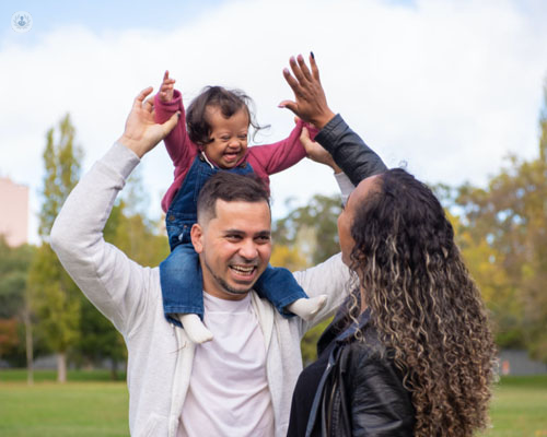 Familia feliz con una bebé con síndrome de down que lleva el padre en hombros - by Top Doctors