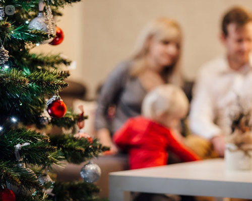 Primer plano de un árbol de Navidad y, en el fondo, una familia con un bebé - Vacaciones de Navidad saludables - by Top Doctors