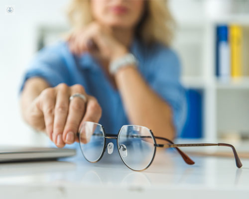 Gafas en primer plano y mujer borrosa en segundo plano, cogiéndolas - cataratas y astigmatismo - by Top Doctors