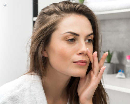 Chica mirándose al espejo tocándose la cara, en actitud de aplicar crema o similar - acné y rosácea - by Top Doctors